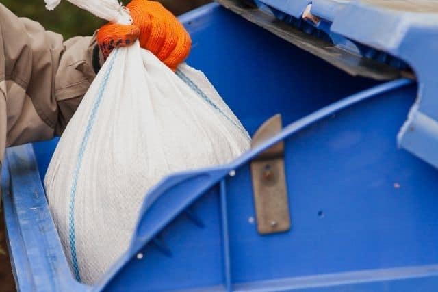 A person placing a double-bagged dead rabbit into an outdoor trash can